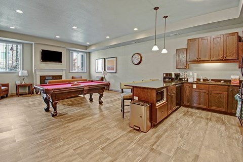 A stylish community lounge here at Williston Garden Apartments with wood-style flooring, recessed lighting, and a welcoming open layout. A pool table anchors the seating area near a wall-mounted television and fireplace, while a full kitchen with wood cabinets, an island, and pendant lighting creates a comfortable space for gatherings and social events.