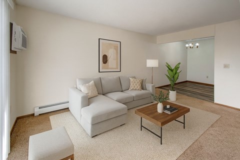A cozy living room here at Windscape Apartments featuring plush carpet, soft neutral walls, and a modern sectional sofa arranged on a textured area rug. A wooden coffee table, simple wall art, and potted greenery add warmth, while an open view into the dining area creates a comfortable, inviting layout.
