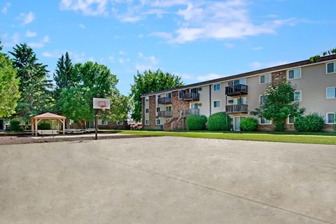 A bright outdoor recreation area here at Windscape Apartments featuring a large open concrete court with a basketball hoop, surrounded by green lawns, mature trees, and nearby apartment buildings with private balconies, creating a welcoming community space for residents to enjoy outdoor activities and social gatherings.
