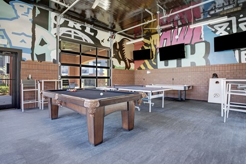 A lively game room here at 71 at Tulsa Hills featuring a pool table, ping pong table, cornhole boards, mounted TVs, and bold mural artwork, creating a fun and social space for residents to relax and gather.