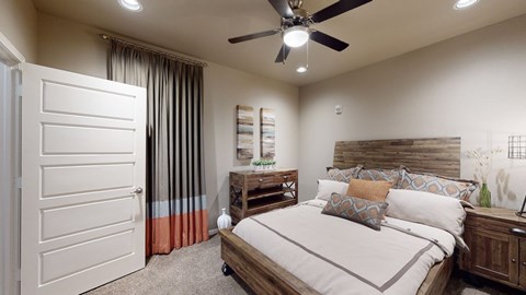 A warm bedroom here at 98 Apartments with soft carpet, a wood-accent bed frame, neutral bedding with patterned pillows, a ceiling fan, and floor-length curtains, creating a cozy and restful retreat.