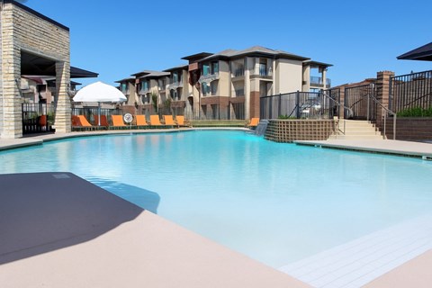 Here at 98 Apartments, a resort-style outdoor pool with clear blue water, modern stone accents, bright orange lounge chairs, and surrounding apartment buildings that create a relaxing and upscale atmosphere.