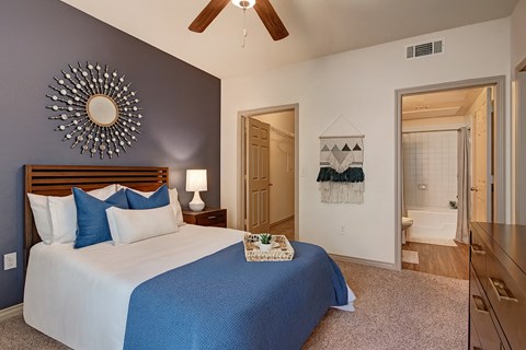 A cozy bedroom here at Aviare Arts District with soft carpet, a blue accent wall, a comfortable bed with neutral and blue bedding, a ceiling fan, and an attached bathroom visible, creating a calm and inviting retreat.