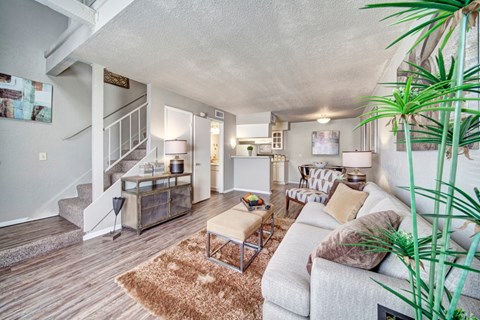 A cozy living room here at Brookwood Village with wood-style flooring, soft neutral furnishings, and warm lighting, featuring an open layout with a staircase, a small dining area, and a connected kitchen that creates an inviting and functional living space.