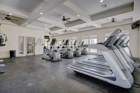 A fitness center here at Liberty Pointe featuring treadmills, stationary bikes, and elliptical machines arranged across an open workout floor. The space has bright recessed lighting, ceiling fans, and multiple windows that bring in natural light for a clean, energized atmosphere.