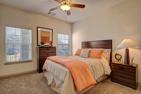 Here at Liberty Pointe, a bright bedroom with warm neutral walls and soft carpet, featuring a wooden bed frame styled with a white bedspread and standout coral bedding accents. Matching wood nightstand and dresser add warmth, while a table lamp, framed wall art, and natural light from two windows create a clean, comfortable feel.
