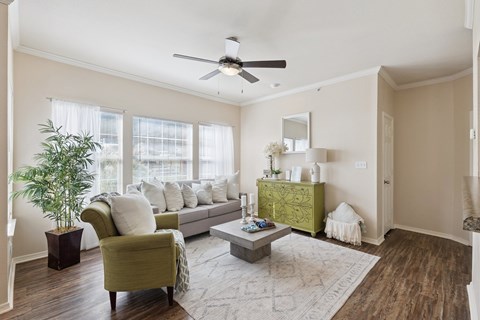 A living room here at Lincoln at Central Park featuring a bright, modern space with a light gray sofa, a green accent chair, and a green decorative dresser that adds a standout pop of color. Large windows with white curtains bring in natural light, while a light area rug, square coffee table, and indoor plant create a clean, comfortable feel.