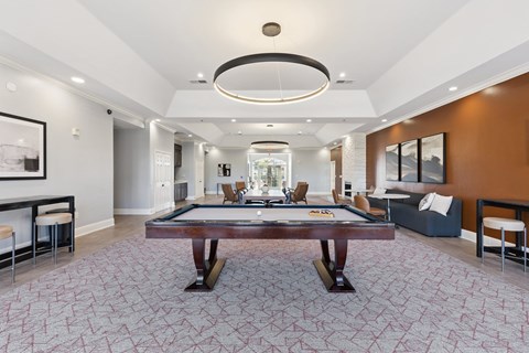 A modern resident lounge here at Lincoln at Central Park with plush carpet flooring, a pool table centerpiece, and stylish circular ceiling lighting. A warm accent wall, comfortable seating, and dining space create a welcoming, upscale social atmosphere.