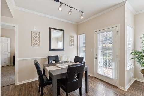 A dining area here at Lincoln at Central Park featuring a dark wood dining table paired with four black upholstered chairs, set on warm wood-style flooring. Natural light enters through a glass patio door and side window with white blinds, while a wall mirror, decorative wall panels, and a simple candle centerpiece create a clean, welcoming, and comfortable space.