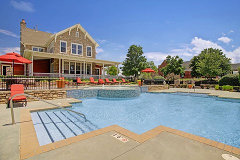 A resort-style pool here at Memorial Creek featuring clear blue water, stone accents, red lounge chairs with umbrellas, wide deck seating, and a bright clubhouse backdrop under a sunny sky.