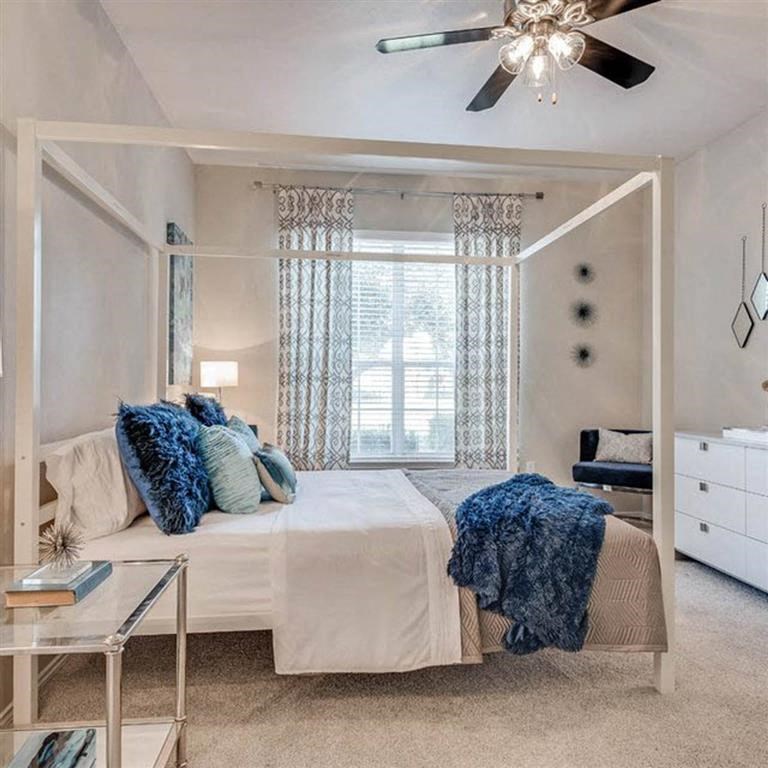 Modern bedroom here at Sycamore Farms Apartment Homes with a white canopy bed, decorated with blue pillows and a throw. Elegant curtains, ceiling fan, and dresser add sophistication.