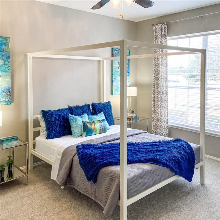Bright bedroom here at Sycamore Farms Apartment Homes with a modern canopy bed, adorned with vibrant blue pillows and throw, abstract blue artwork, and a large window with patterned curtains.