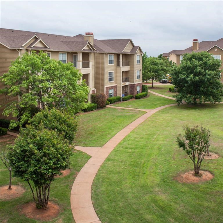 Sycamore Farms Apartment Homes with beige buildings and brown roofs surrounded by lush green grass and trees. Curved pathways lead through a peaceful, manicured setting.