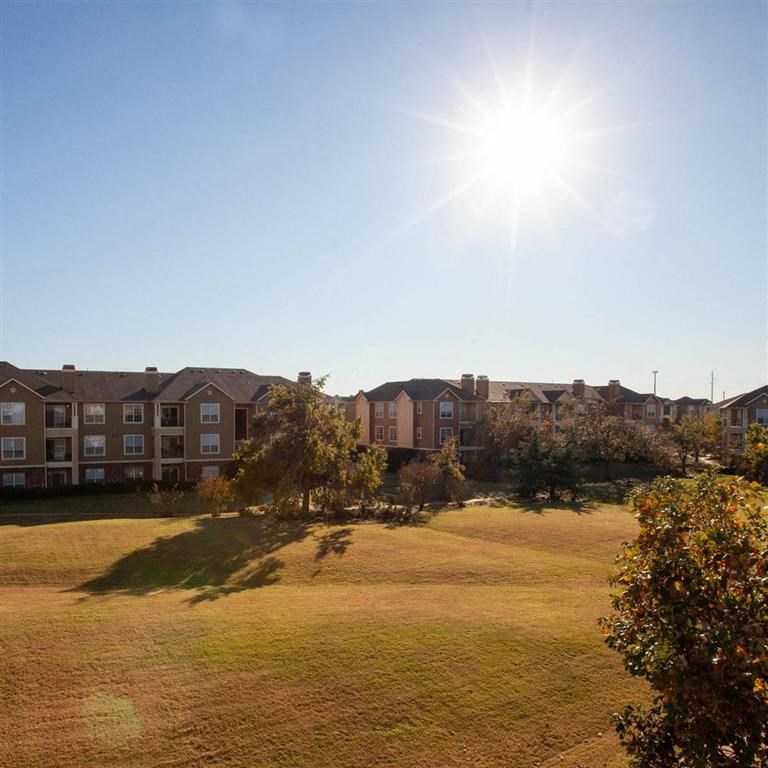 Sunny day view of Sycamore Farms Apartment Homes with brown apartment buildings and a grassy field dotted with trees, conveying a peaceful autumn atmosphere.