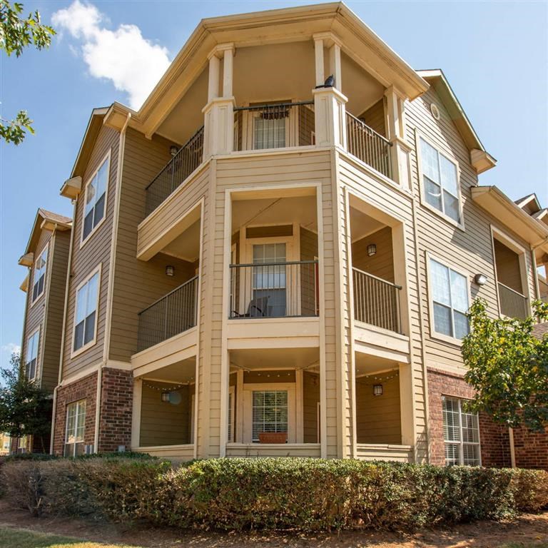 Three-story Sycamore Farms Apartment Homes building with balconies, set against a clear blue sky. Surrounded by green trees and neatly trimmed bushes. Sunny and serene atmosphere.