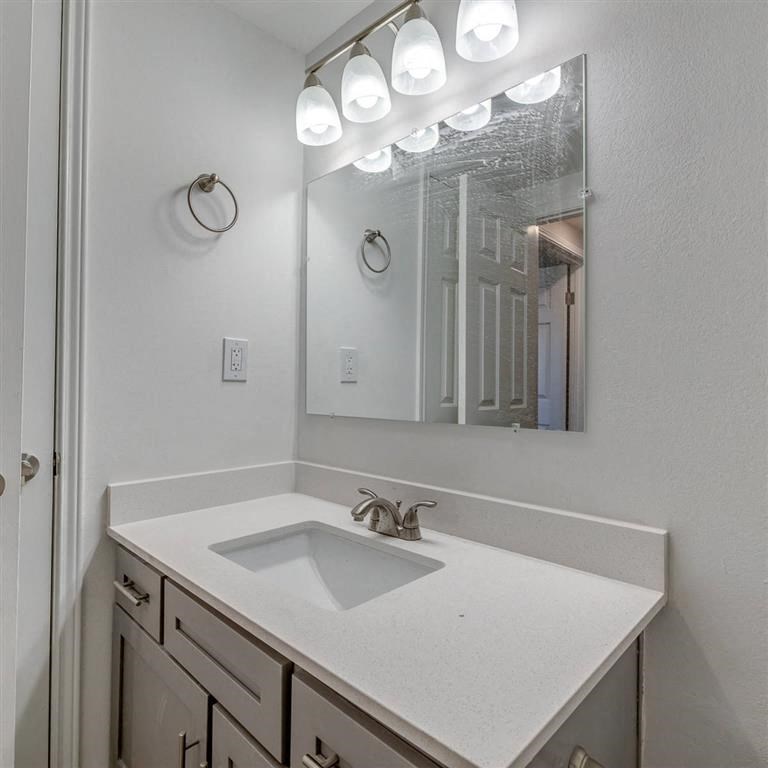 Small bathroom here at Sycamore Farms Apartment Homes with a sleek white countertop, undermount sink, and silver faucet. Above, a broad mirror reflects a closed door, framed by bright vanity lights.