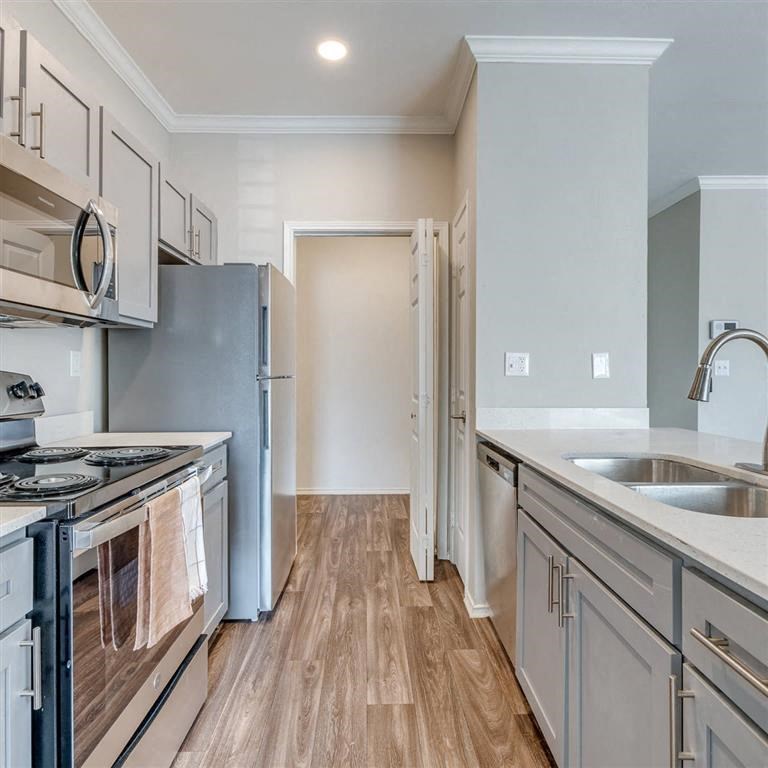 Modern kitchen here at Sycamore Farms Apartment Homes with stainless steel appliances, light gray cabinetry, and wood-style flooring. A double sink is set in the white quartz countertop.