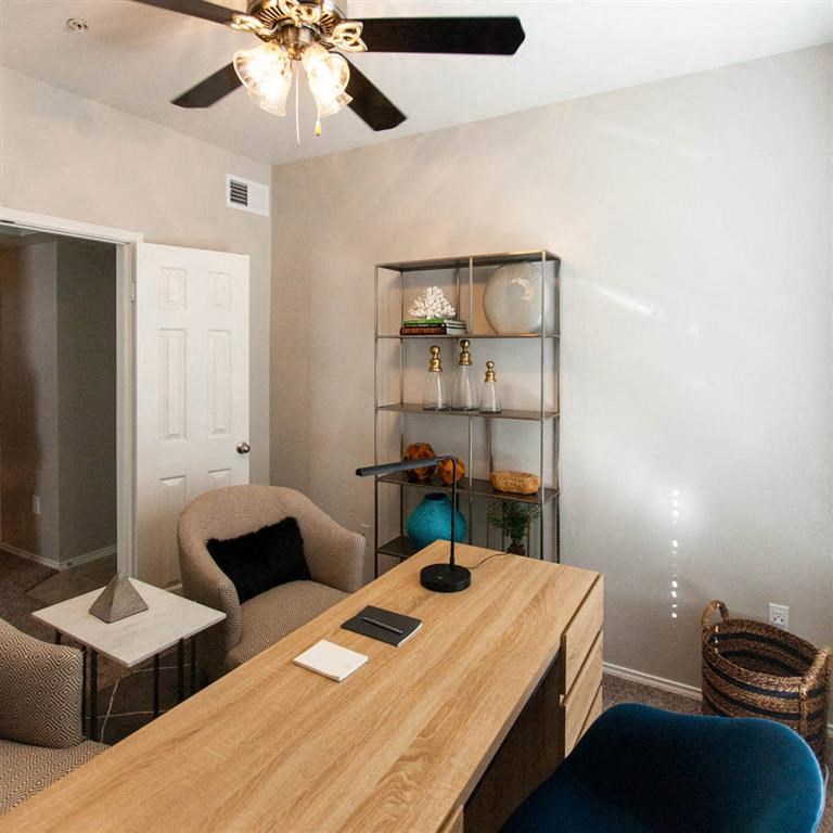 Cozy home office here at Sycamore Farms Apartment Homes with a wooden desk, modern lamp, beige armchair, and shelves displaying decorative items. Ceiling fan cast soft light. Calm ambiance.