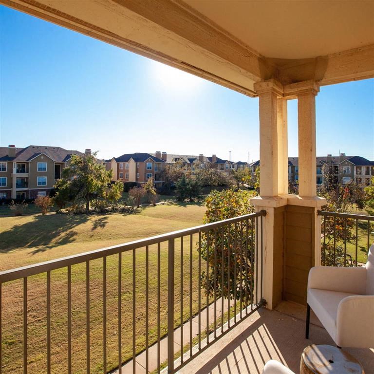Balcony here at Sycamore Farms Apartment Homes with white chair overlooks a sunny grassy field and apartment buildings. The bright blue sky creates a serene, inviting atmosphere.