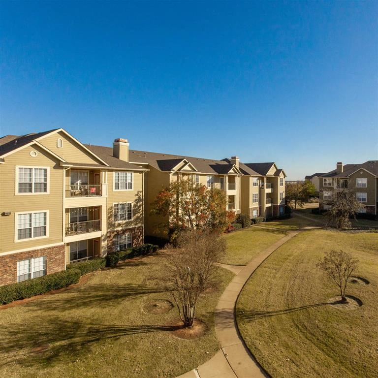 Sunny, spacious Sycamore Farms Apartment Homes with beige buildings and white windows. A curved path winds through a green lawn, creating a peaceful atmosphere.