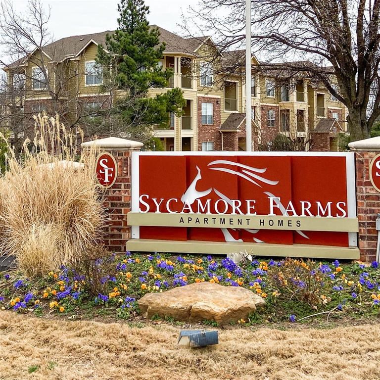 Entrance sign for Sycamore Farms Apartment Homes with landscaping. Background features a multi-story brick and beige building, trees, and a clear sky.