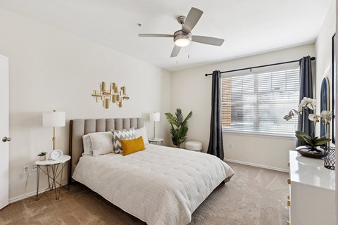 A bedroom here at Quail Landing with soft carpet, a light upholstered bed, white bedding with a mustard accent pillow, matching side tables with lamps, a ceiling fan, and natural light through wide windows with dark curtains.