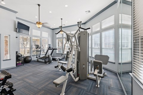 A fitness center here at Quail Landing with modern strength machines, cardio equipment, blue patterned flooring, and tall windows that fill the space with bright natural light.