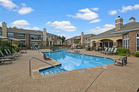 A resort-style pool area here at Quail Landing with clear blue water, tan pool decking, black lounge chairs, shaded seating, and surrounding brick apartment buildings under a bright blue sky.