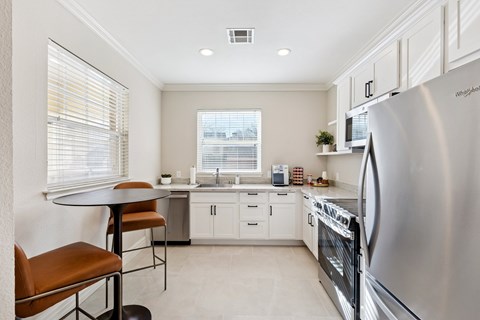 A modern kitchen with a stainless steel refrigerator, a table with two chairs, and a window with blinds.