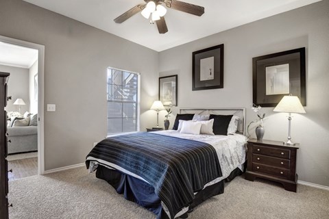 A comfortable bedroom here at Stoneleigh on May with beige carpet, soft gray walls, and a ceiling fan with light. A large bed with neutral bedding is flanked by matching nightstands, while a window with blinds brings in natural light.