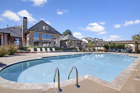 A resort-style swimming pool here at Stoneleigh on May with clear blue water, poolside lounge chairs, and surrounding greenery. A clubhouse backdrop and open sky create a relaxing, community-focused outdoor setting.