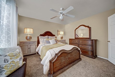 A spacious bedroom here at Summit Point with plush carpet, soft neutral walls, and a ceiling fan with light. A large bed with classic wood furniture and warm lighting creates a comfortable, inviting retreat.