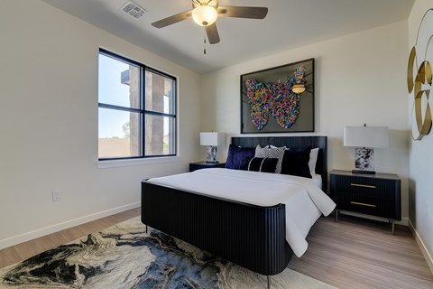 A bedroom here at Trailwinds with a black bed frame, white bedding, dark nightstands, colorful butterfly wall art, ceiling fan, large window, and soft patterned rug creating a bright, modern feel.