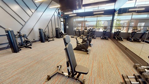 A fitness center here at Trailwinds with modern cardio and strength machines, patterned flooring with subtle orange accents, mirrored walls, high ceilings, and large windows bringing in natural light.