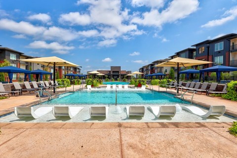 A resort-style pool here at Trailwinds with a shallow lounging ledge, white in-water chaise loungers, blue umbrellas, surrounding lounge chairs, and landscaped courtyards framed by modern apartment buildings under a bright blue sky.