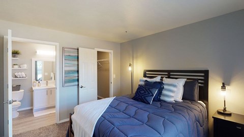 A bedroom here at Villas at Countryside with blue bedding on a black bed frame, soft gray walls, warm bedside lighting, and an open view into a bright bathroom and closet creating a calm, welcoming feel.