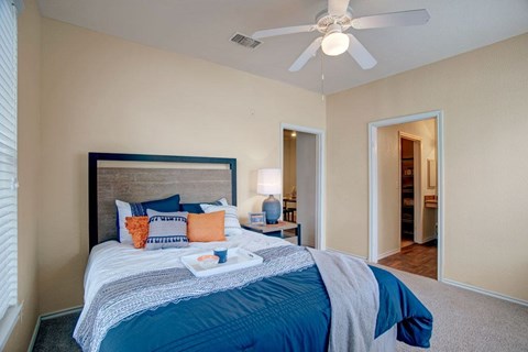 A bedroom here at Woodland Park with a beige color palette, white ceiling fan, and carpeted floor, featuring a wood-tone headboard bed with blue and orange accent pillows, a bedside lamp, and open doorways to a closet and bath area.