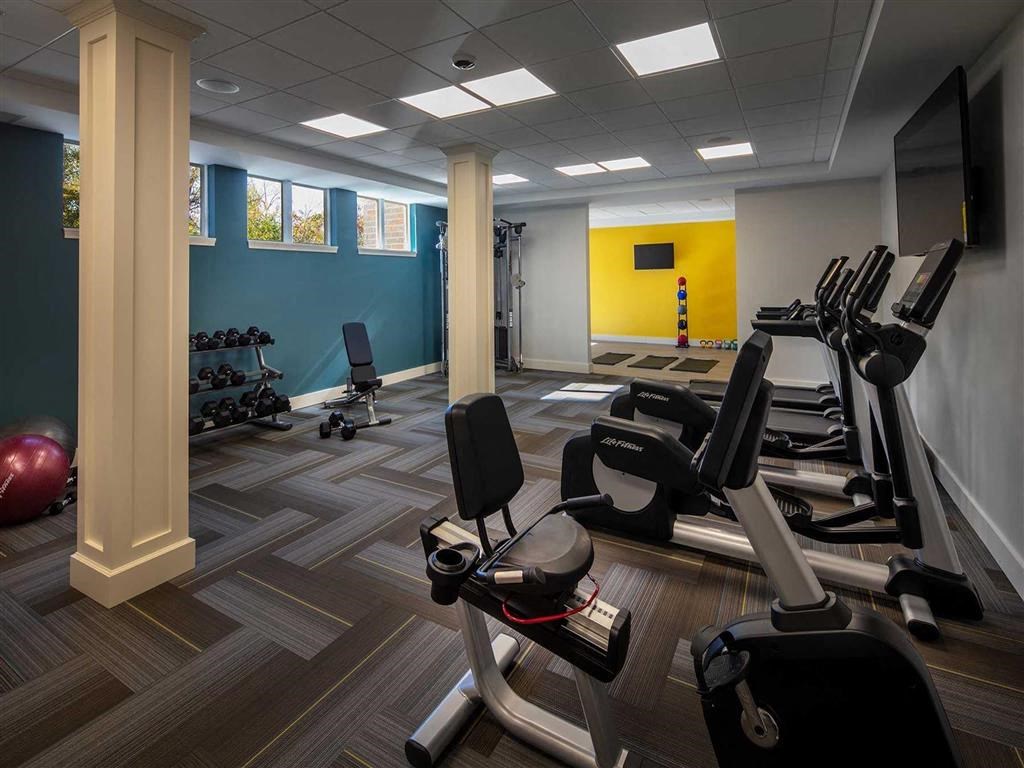 Compact gym with stationary bikes, a row of dumbbells, exercise balls, and a weight machine at the Oaks of Shorewood Apartments. Walls are blue and yellow, creating a vibrant, energetic tone.