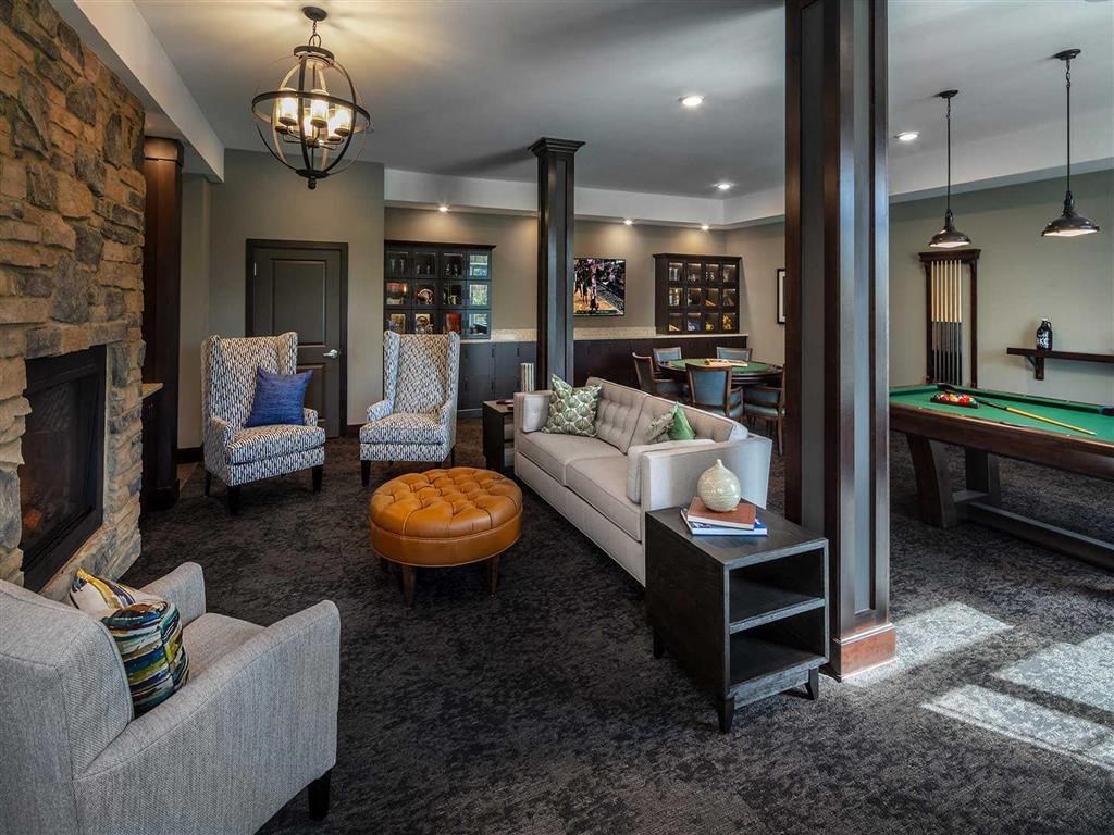 Cozy resident lounge with plush armchairs, a tan ottoman, and a beige sofa, accented with pillows. Features a stone fireplace and a pool table, creating a warm, inviting atmosphere. The Oaks of Shorewood Apartments.