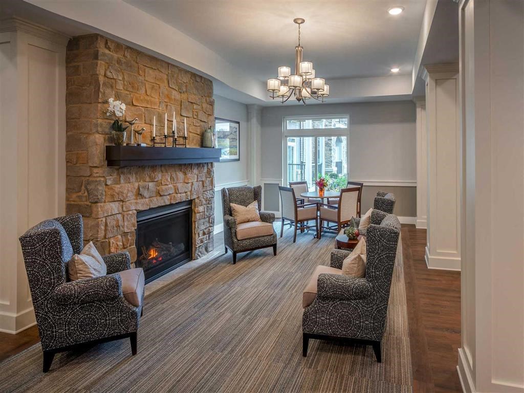 Cozy living room at the Oaks of Shorewood Apartments with a stone fireplace, flanked by cushioned armchairs. A chandelier hangs over a dining table near a large window, creating a warm ambiance.