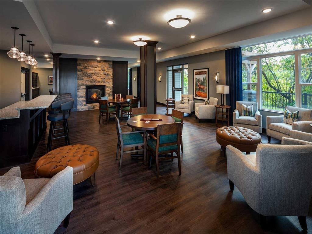 Cozy lounge at the Oaks of Shorewood Apartments with dark wood floors, plush chairs, and a stone fireplace. Large windows offer a view of greenery, creating a warm, inviting atmosphere.