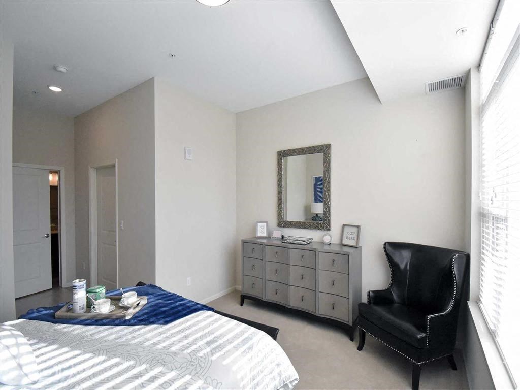 Bright bedroom at the Oaks of Shorewood Apartments with a bed, blue throw, and tray of cups. Gray dresser with mirror, black armchair, and soft lighting create a cozy, modern feel.