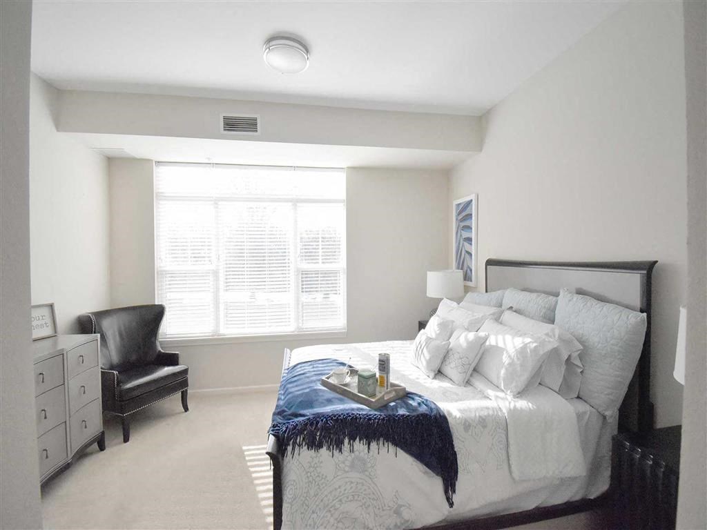 Bright bedroom at the Oaks of Shorewood Apartments with a large window, featuring a neatly made bed with a blue throw, a black armchair, and a dresser. The tone is tranquil and inviting.