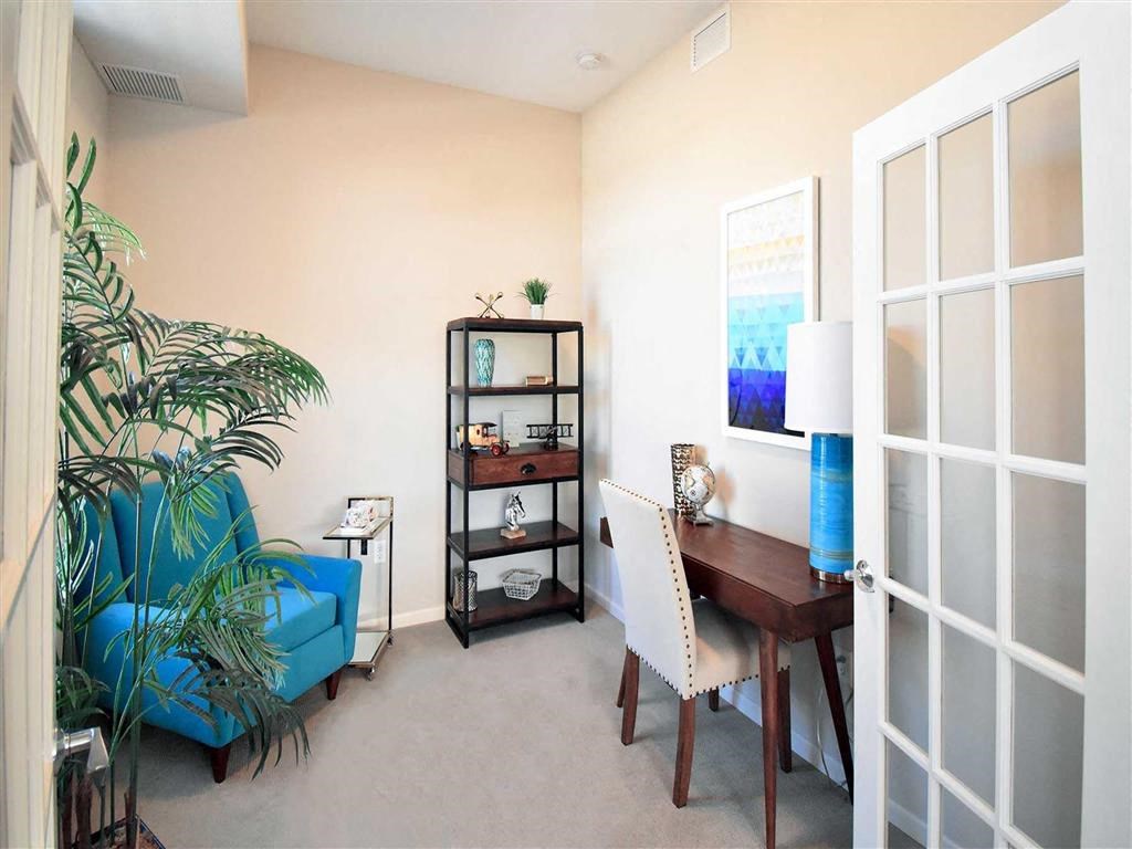 Cozy office at the Oaks of Shorewood Apartments with a blue armchair, potted plant, and shelf against cream walls. A wooden desk has a white chair, lamp, and abstract blue artwork.