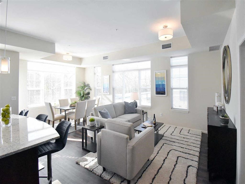 Bright, modern living space at the Oaks of Shorewood Apartments with a gray sofa, textured rug, dining table, and barstools. Large windows, soft lighting, and a cozy, inviting atmosphere.