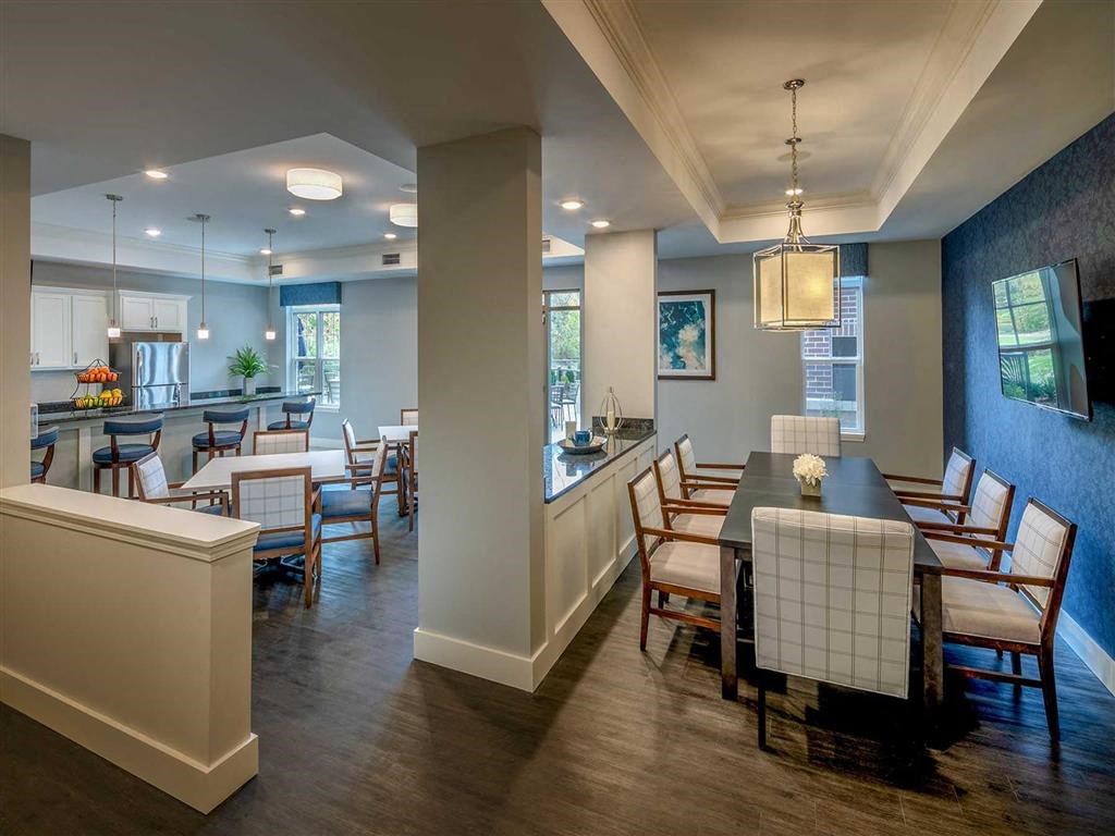 Resident lounge at the Oaks of Shorewood Apartments with modern kitchen and dining area with wood flooring, white cabinetry, and blue accents. A long dining table seats eight. Bright, welcoming atmosphere.