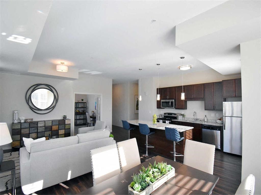 Modern open-concept living space at the Oaks of Shorewood Apartments with a cozy ambiance. Features a sleek kitchen with dark cabinets, island seating, and a stylish living area with a gray sofa.