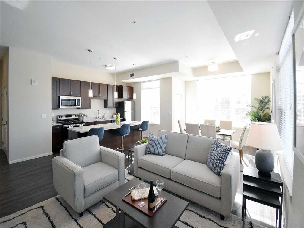 Modern open-concept living area at the Oaks of Shorewood Apartments with a cozy gray sofa, armchair, and coffee table. Bright kitchen and dining space in the background with natural light.