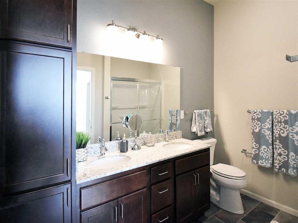 Modern bathroom at the Oaks of Shorewood Apartments with dark wood cabinets, dual sinks, and granite countertop. Wall-mounted lights illuminate the space. Stylish towels hang near the toilet.