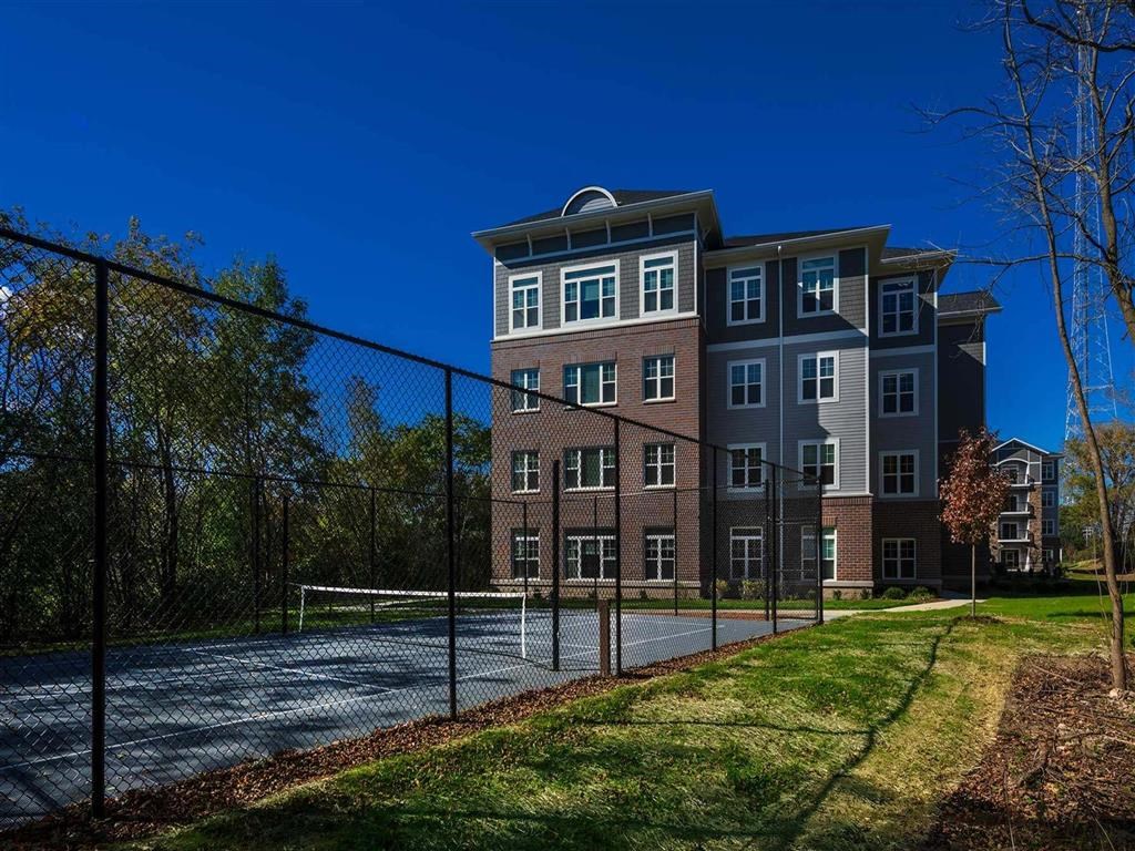 Four-story brick building with white-trimmed windows beside a fenced tennis court. Surrounded by trees under a clear blue sky, creating a peaceful atmosphere. The Oaks of Shorewood Apartments.